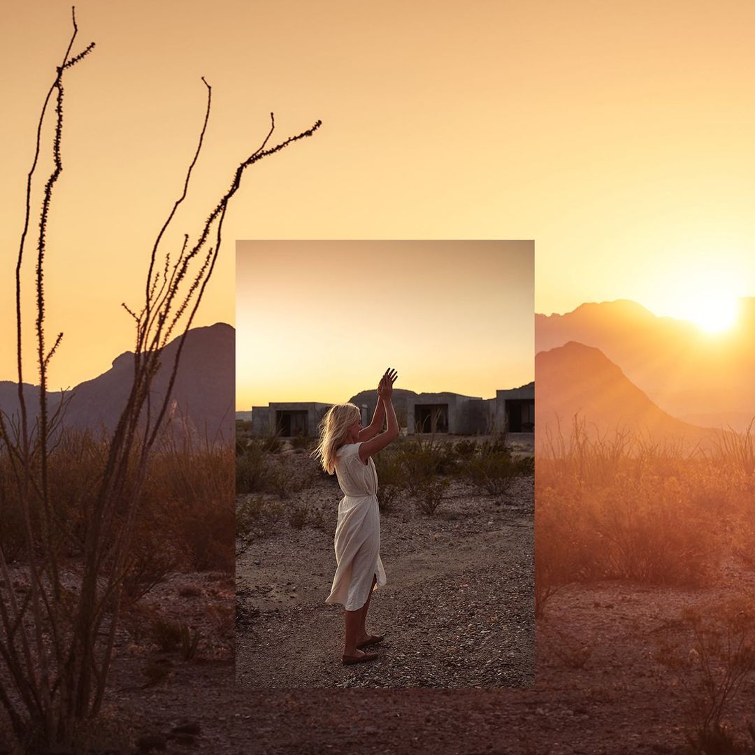 Woman in white dress raises arms in desert landscape with mountains and a golden sunset, framed by desert flora and modern architecture.