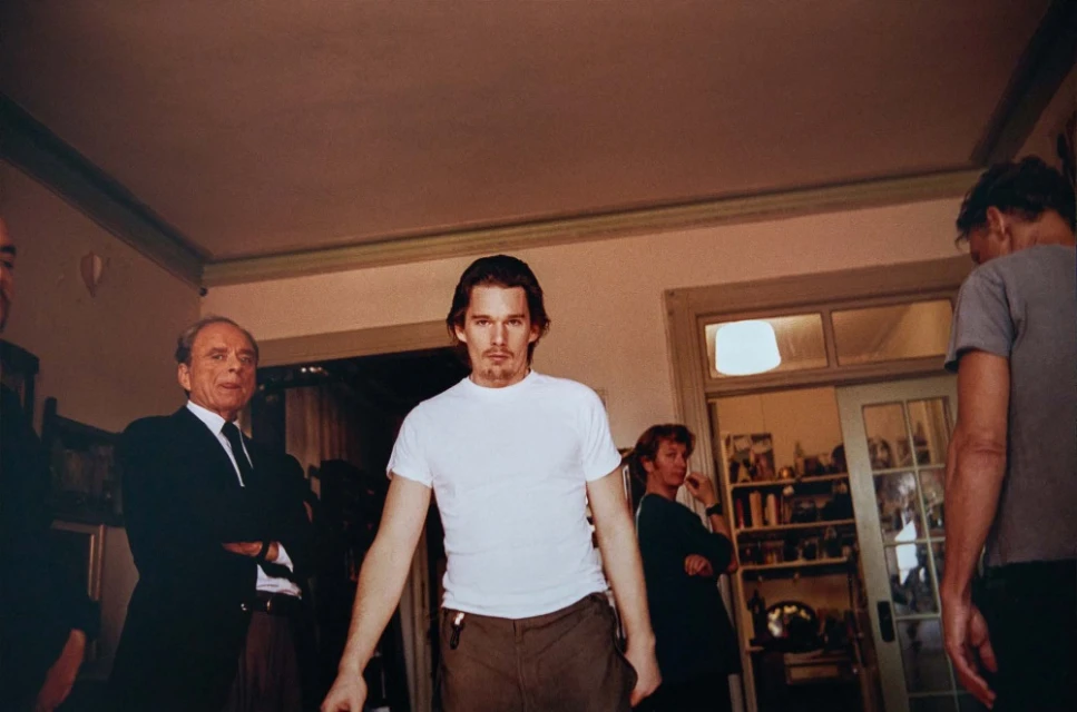 Ethan Hawke stands in a white t-shirt with a group of people in the background, inside a room with a door and window.