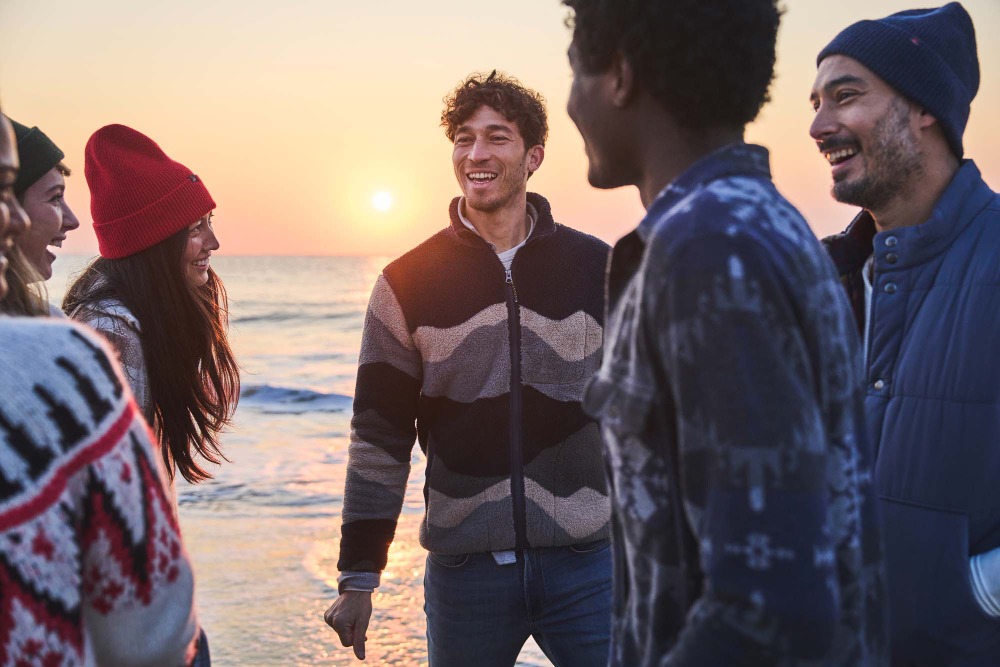 A group of six people stand on a beach at sunset, smiling and talking. The group is diverse, with a mix of men and women.