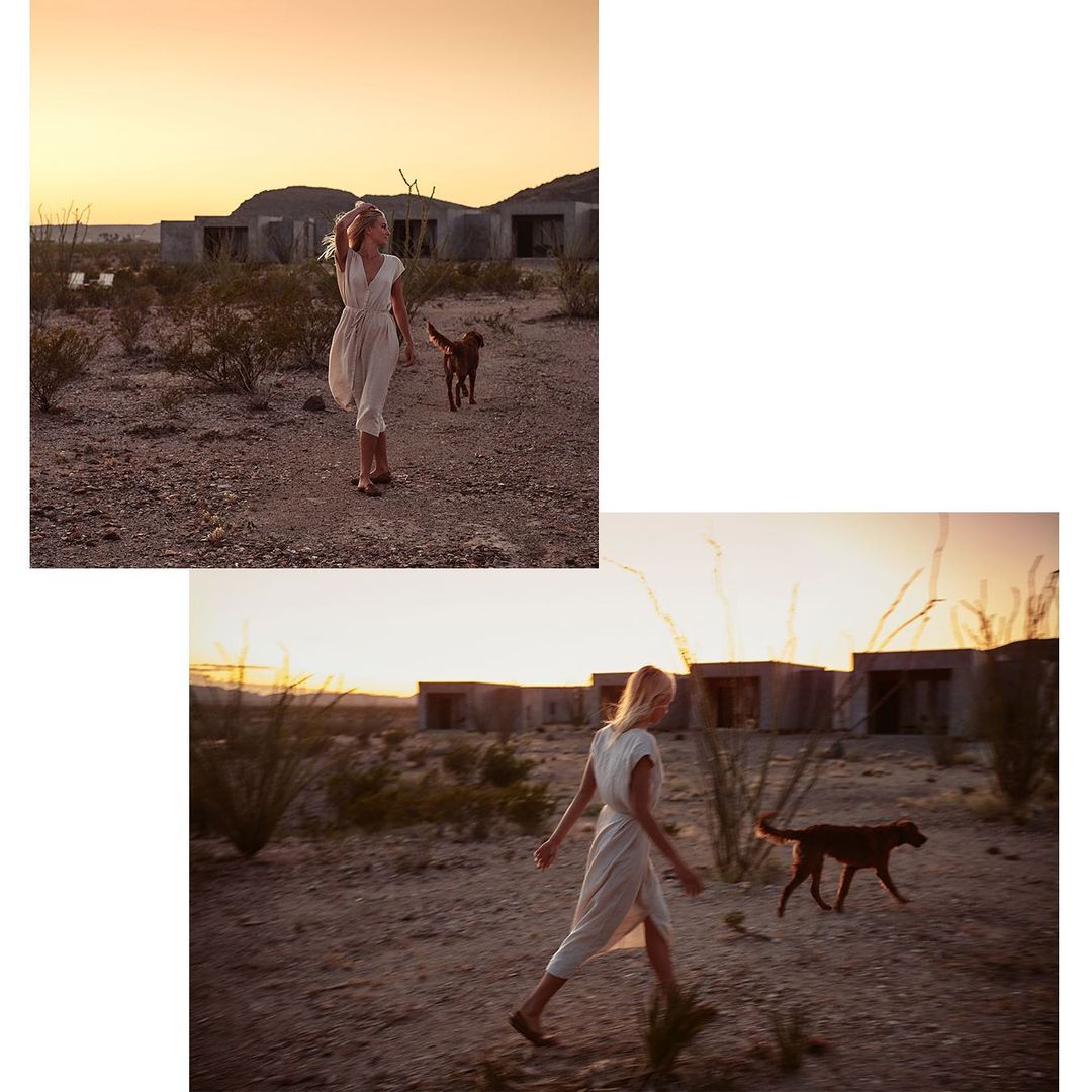 A woman in a dress walking with a brown dog in a desert landscape at sunset with building ruins in the distance.