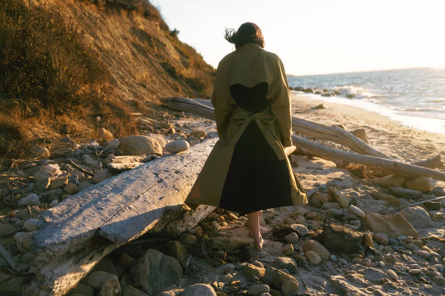 Woman in a green and black dress walking barefoot on a rocky beach toward the ocean, with a grassy bluff in the background.