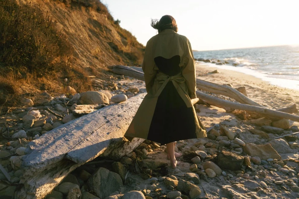 Woman in a green and black dress walking barefoot on a rocky beach toward the ocean, with a grassy bluff in the background.