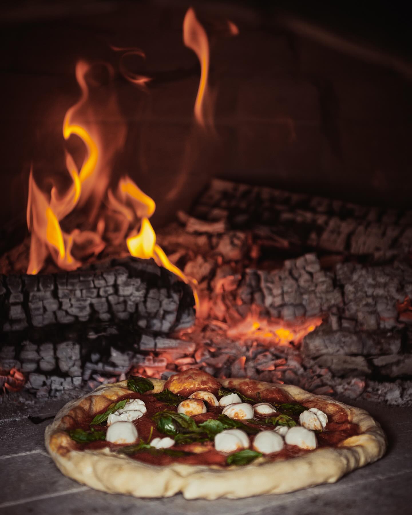 Overhead shot of Neapolitan pizza in wood-fired oven with melted mozzarella, tomato sauce, and basil leaves near burning logs.
