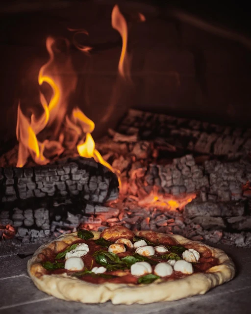 Overhead shot of Neapolitan pizza in wood-fired oven with melted mozzarella, tomato sauce, and basil leaves near burning logs.