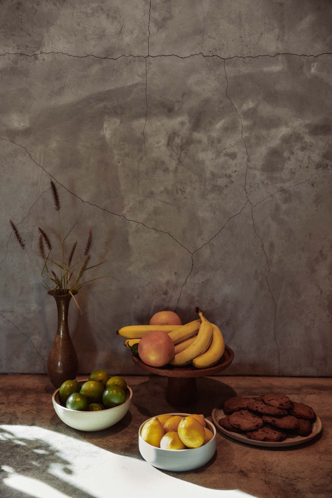 Still life with bananas, apples, citrus, and cookies arranged on a counter against a grey concrete wall, highlighted by shadows.