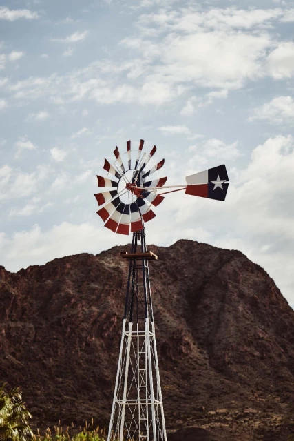 Outdoor scene of a Texas-flag-themed windmill standing in front of a hill under a bright, cloudy sky in the daytime.