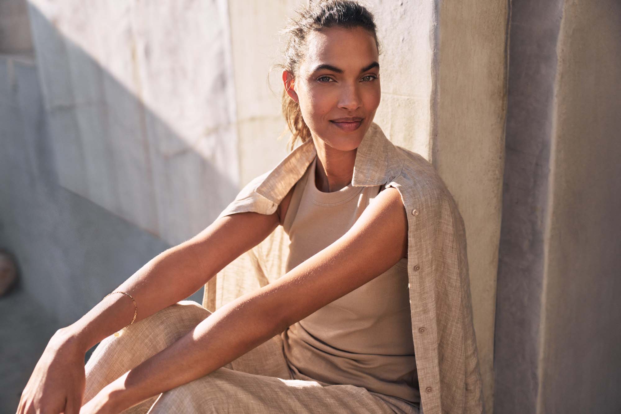 Brunette woman sits against wall wearing beige linen shirt and pants, giving warm smile in bright sunlight. Natural beauty and style.