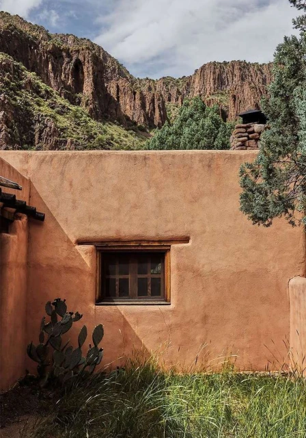 Adobe building with a window and a cactus in front of a rocky mountain landscape with green trees and grass in the foreground.