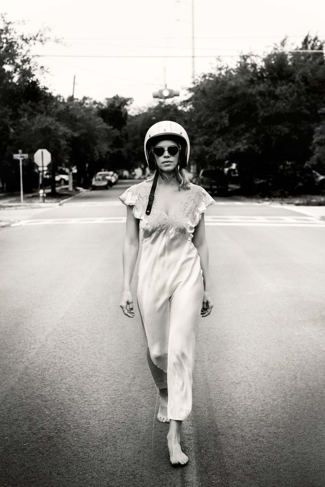 Black and white portrait of a woman with sunglasses and a helmet walking barefoot on a city street with trees in the background.