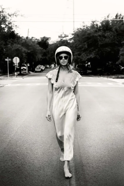 Black and white portrait of a woman with sunglasses and a helmet walking barefoot on a city street with trees in the background.