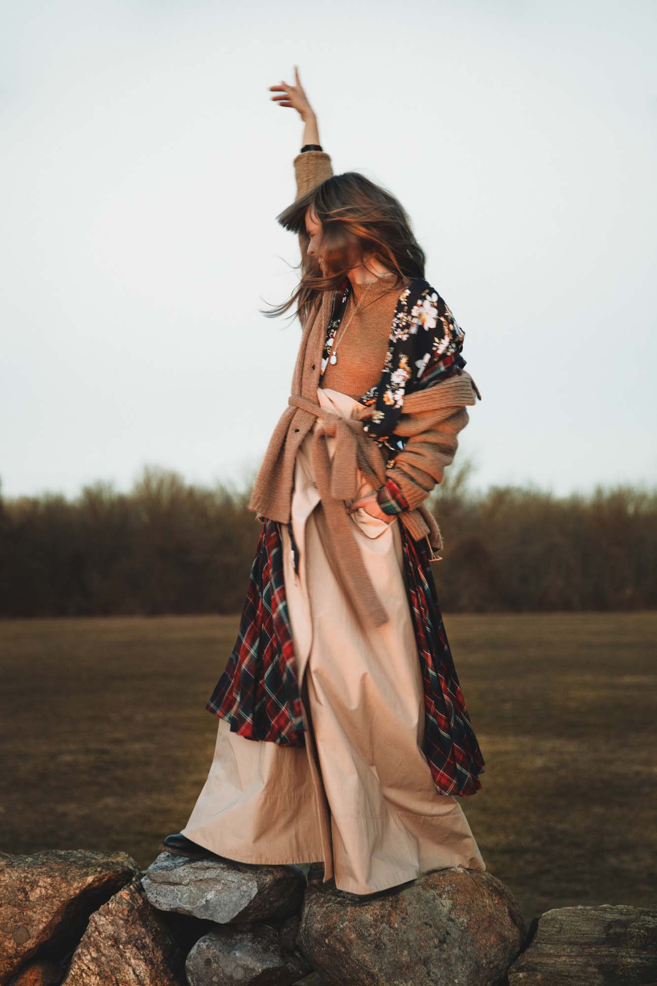 Woman in a belted camel cardigan standing on rock wall, lifting arm against an open grassy field.