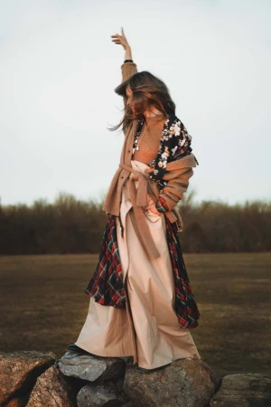 Woman in a belted camel cardigan standing on rock wall, lifting arm against an open grassy field.