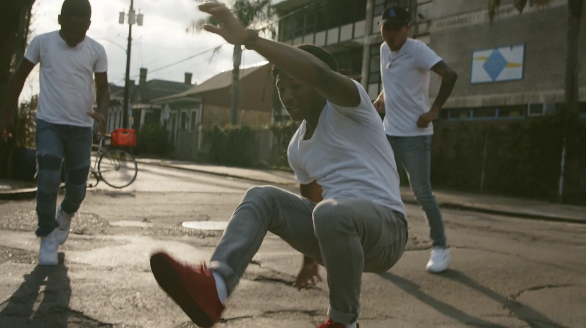 Three young men in casual clothes are on a street, one is in mid-air, and there is a bicycle visible in the background during the day.