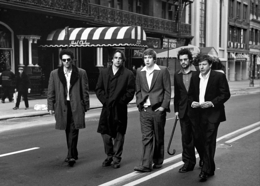 Five men in coats walk across a city street with buildings in the background in a black and white photograph taken during the day.