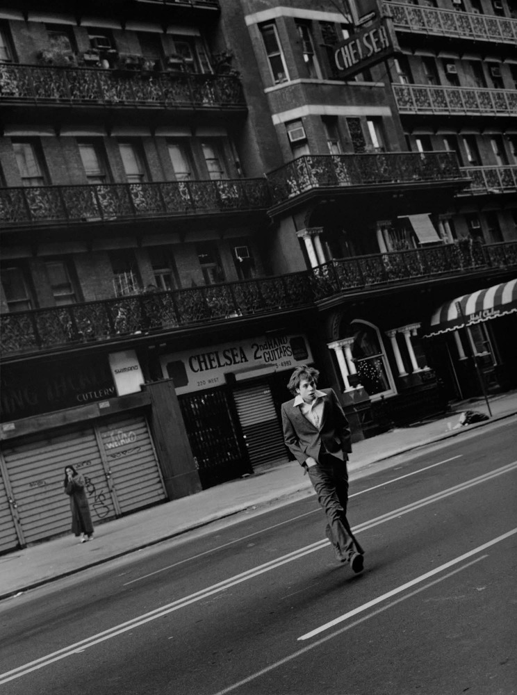 A young man in a suit jacket walking across a New York street in front of the Chelsea building, photographed by Squire Fox.