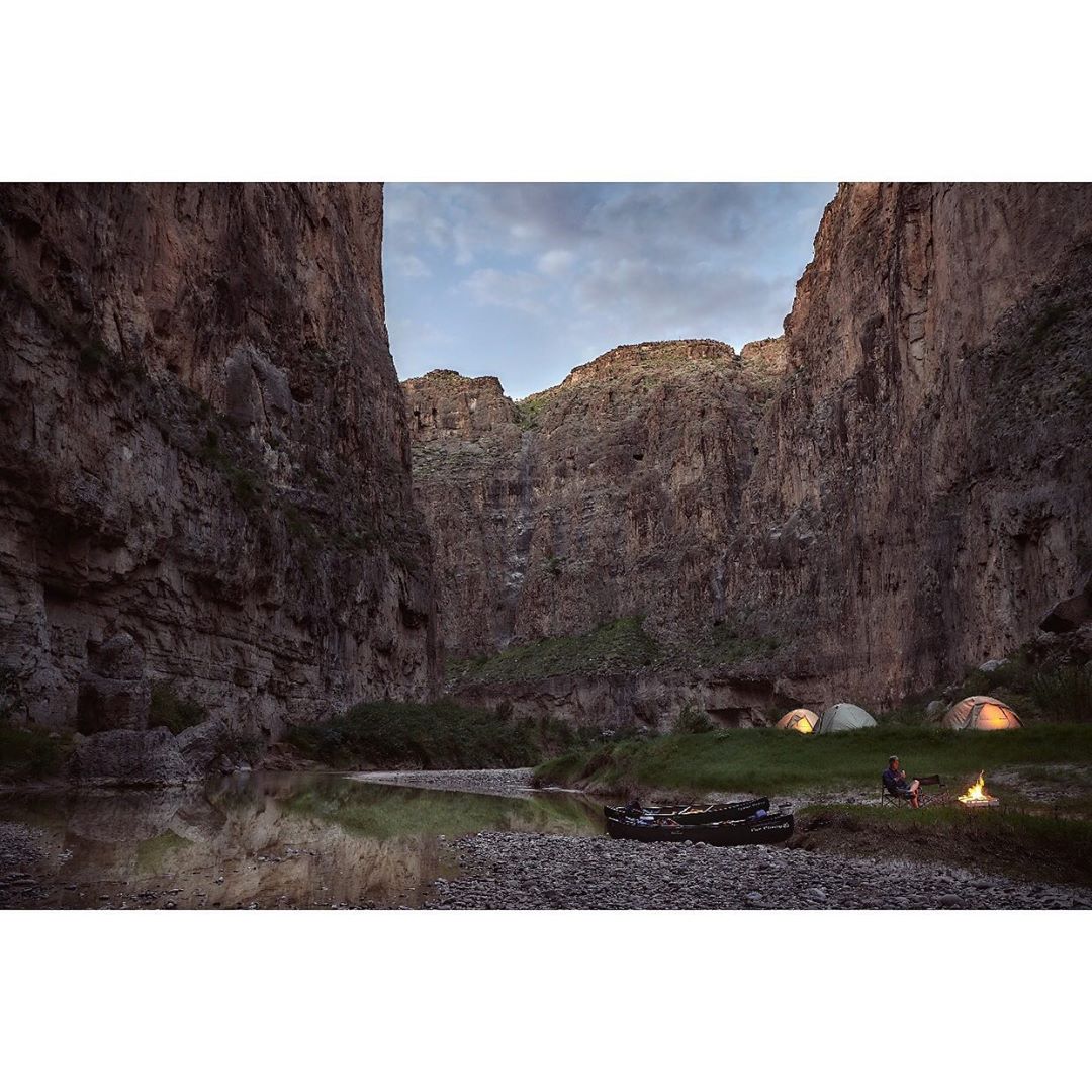 Person sitting by campfire near tents and river in a canyon landscape, lit by the warm glow of the fire and tent lights.