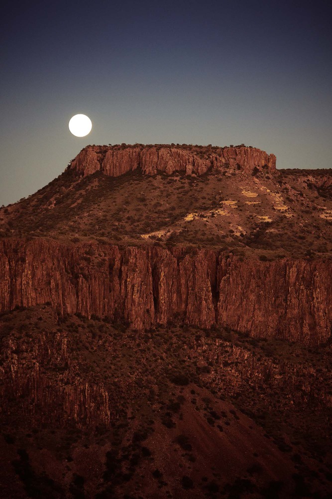 Wide shot of a desert mountain with a full moon in the sky at dusk, displaying nature's grandeur and the tranquil beauty of the landscape.