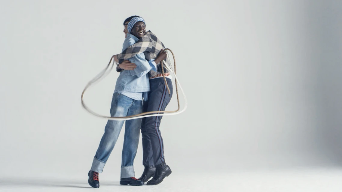 Two people hug in a white studio, wearing jeans and casual tops, a rope lays between them, creating a sense of connection and unity.