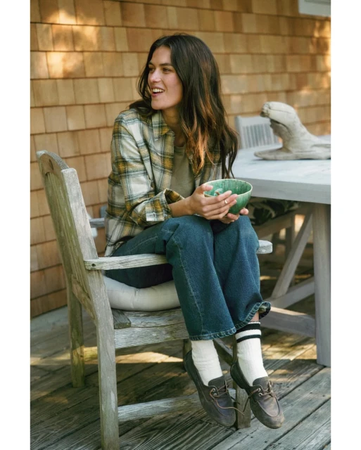 A young woman sits in a wooden chair outside a wood-sided house wearing jeans, a plaid shirt, and brown loafers while holding a green bowl.