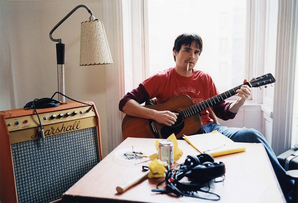 Adult male plays guitar in natural light by window with Marshall amp, table, lamp, cigarettes, headphones, and soda can.