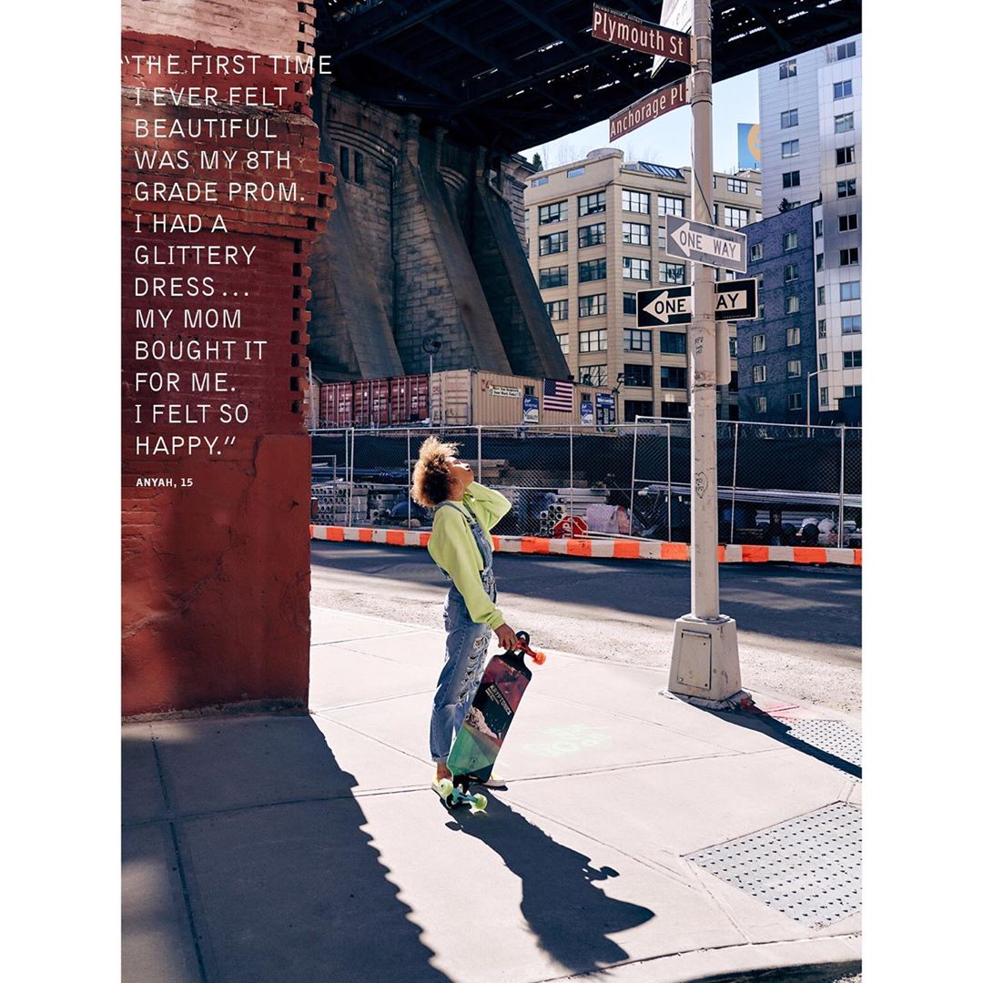 Young adult woman stands with skateboard on a sunny city street under a bridge; street signs and buildings in the background.