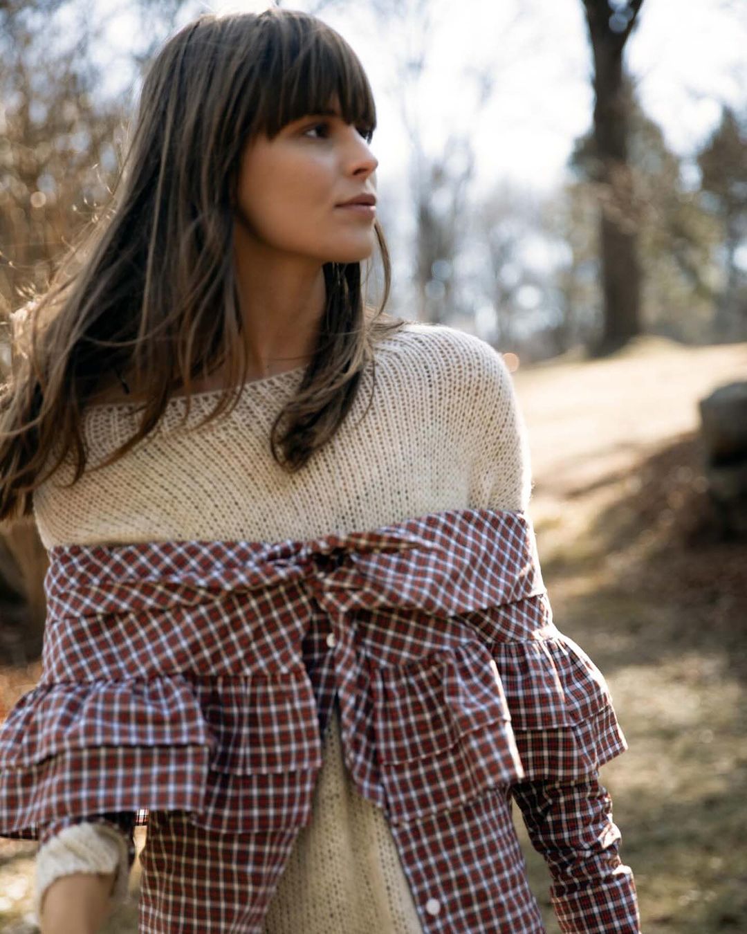 Adult woman with long brown hair poses in natural light wearing knit sweater and brown-and-white checked top in an outdoor setting.