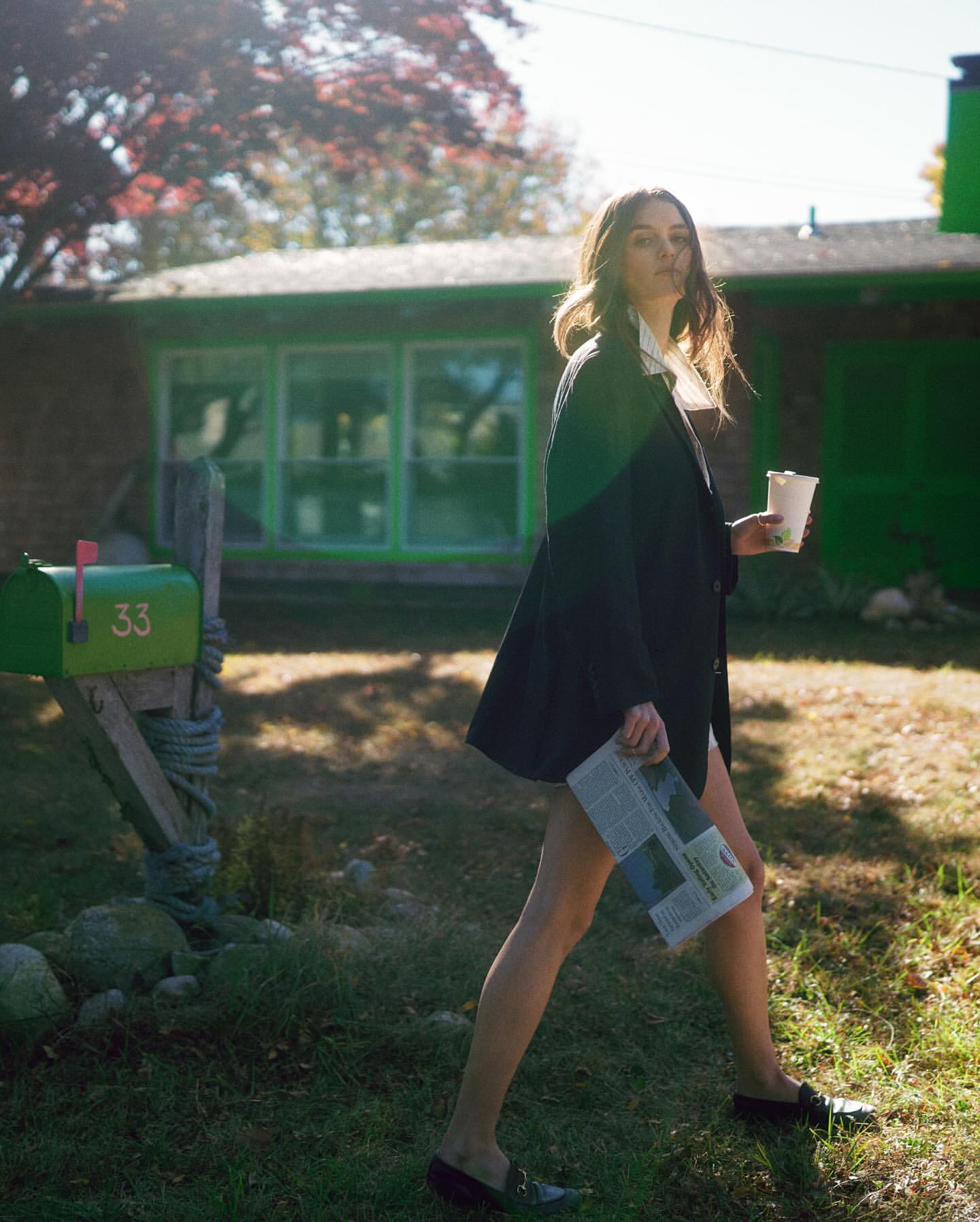 A young woman in a black blazer walks on a grassy lawn with a newspaper and a drink near a green mailbox and a house in daylight.