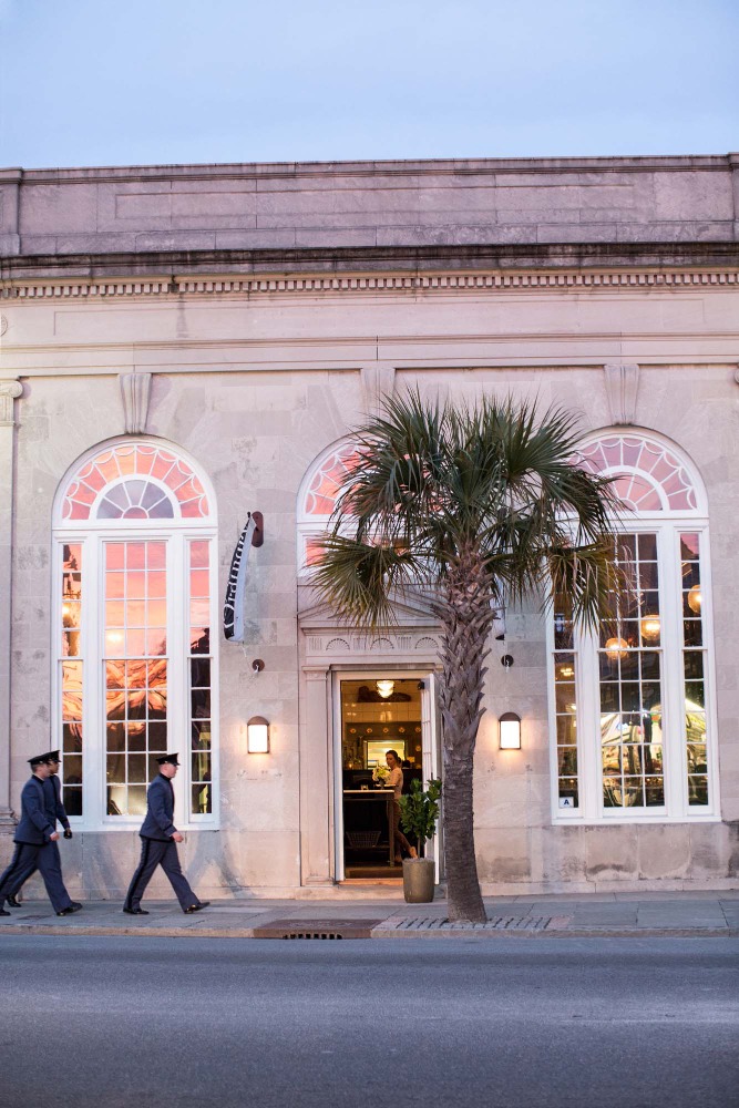 Exterior view of a light stone building with arched windows, a palm tree, and two uniformed people walking along the sidewalk.