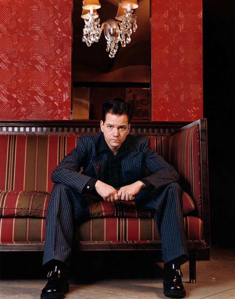 Man in a pinstripe suit sitting on a striped couch indoors, with a chandelier and patterned red wallpaper in the background. Portrait.