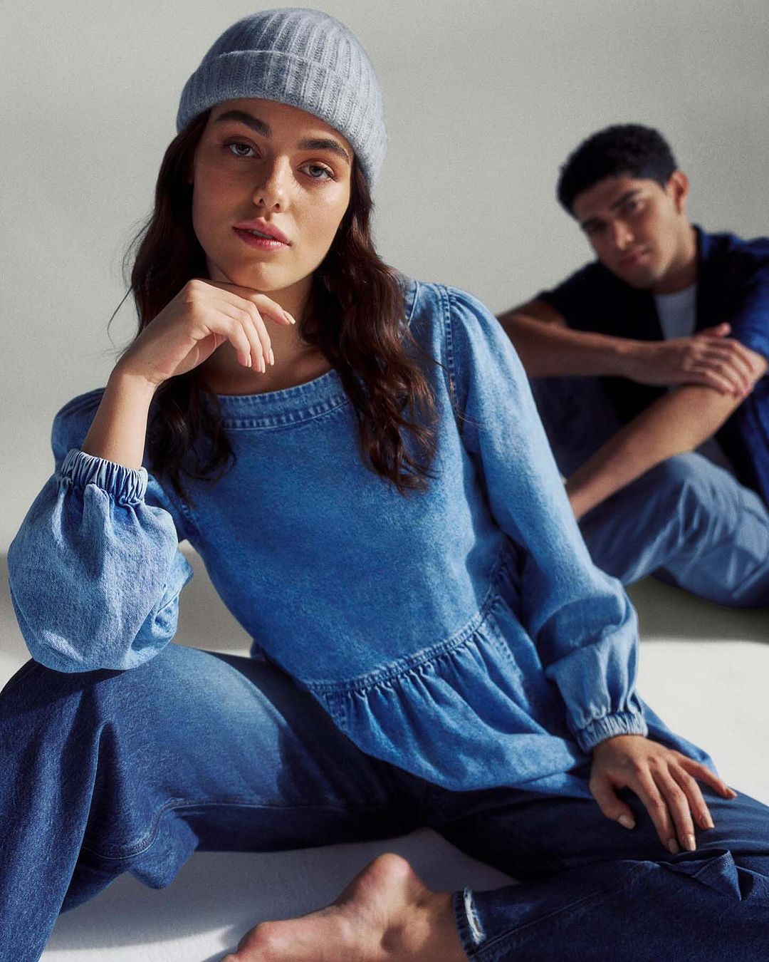 Two fashion models in denim attire pose in a bright studio. A woman in a blue denim top and beanie sits, with a man in a matching shirt behind her.
