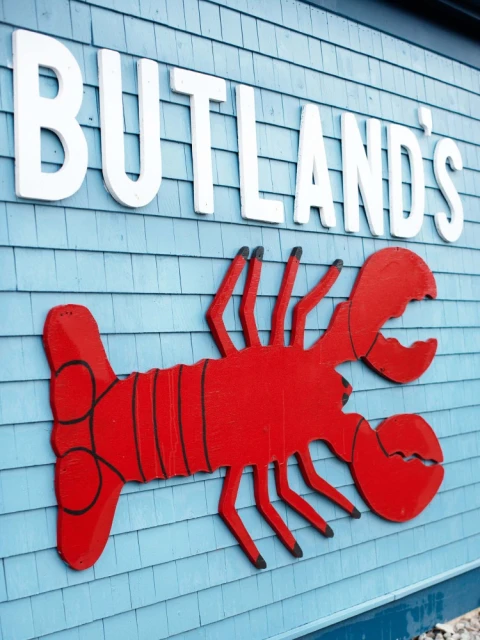 Outdoor sign for Butland's seafood restaurant features a red lobster on light blue siding with white lettering, creating a nautical theme.
