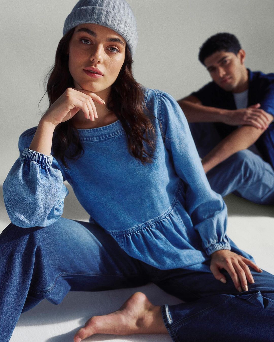 Two people in denim clothing are posing in a studio. A woman with a grey beanie and a denim top in the foreground, a man in the background.