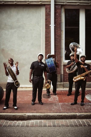 Five Black musicians in uniform standing on a red brick sidewalk outside a brick building, holding brass and percussion instruments.