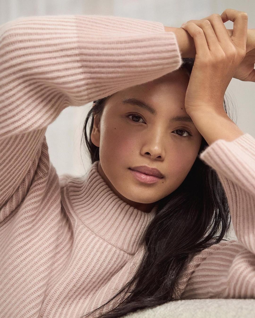 Close-up portrait of a woman wearing a pale pink ribbed turtleneck sweater with her hands raised near her head.