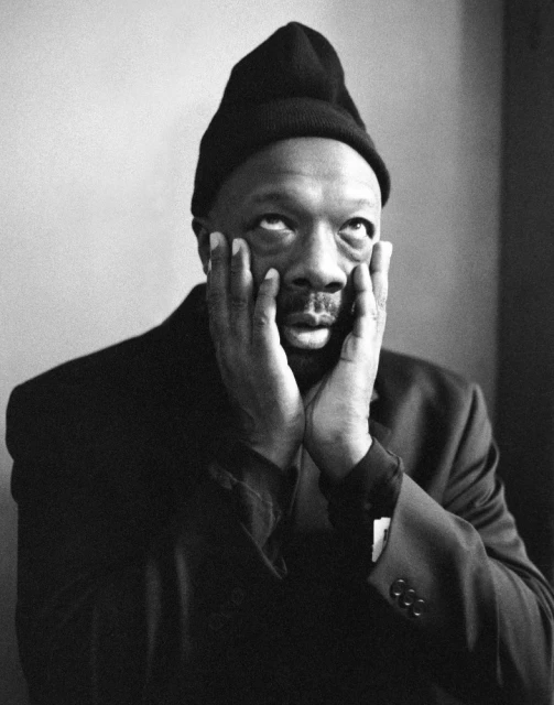 Black and white portrait of an adult male with hands on face, wearing a dark suit jacket and beanie cap, looking upwards.