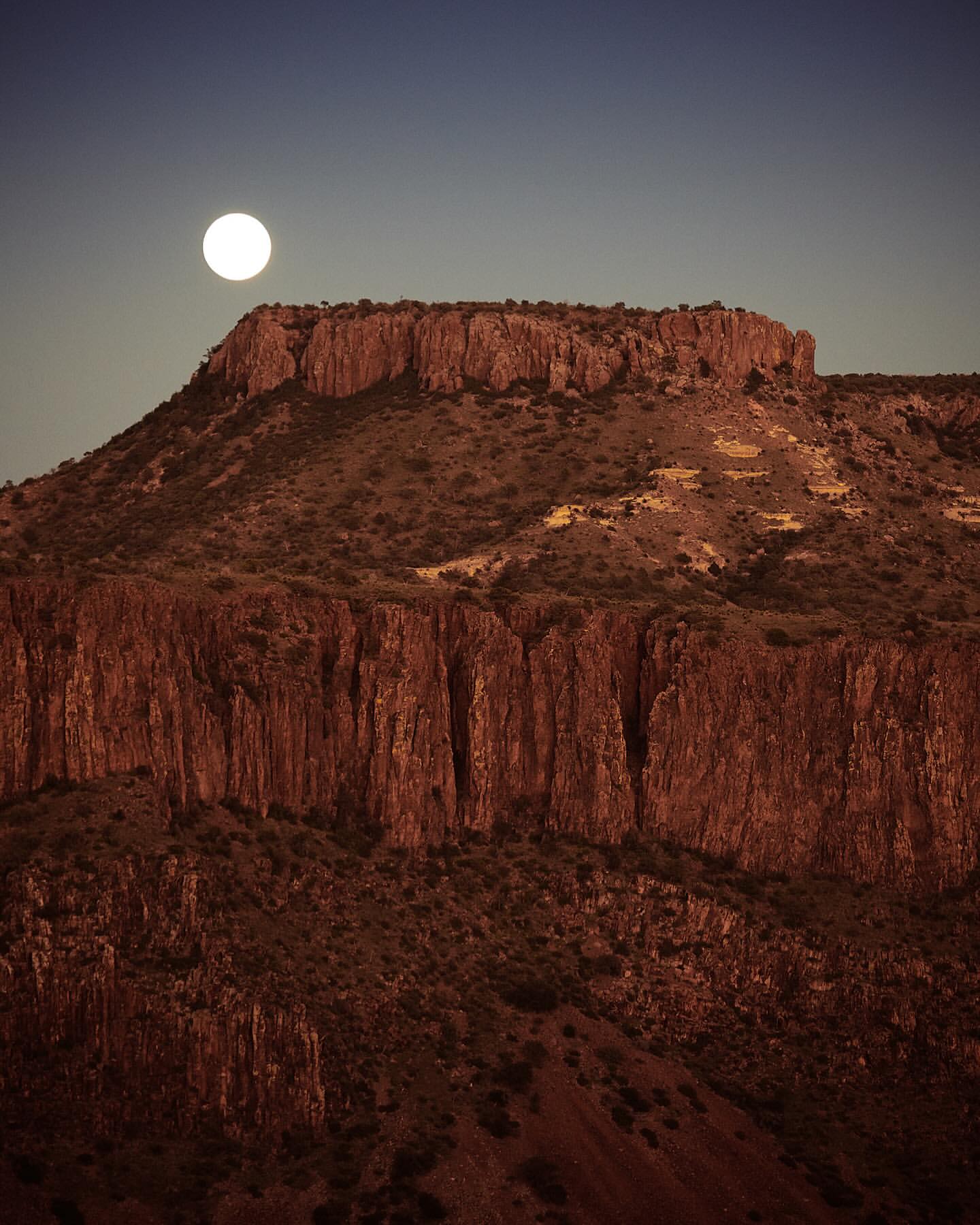 Dramatic landscape photograph showcasing a rugged mountain range under a bright full moon against a gradient sky from dark blue to orange.