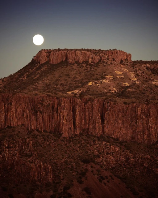 Dramatic landscape photograph showcasing a rugged mountain range under a bright full moon against a gradient sky from dark blue to orange.
