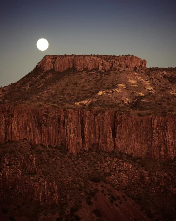 Dramatic landscape photograph showcasing a rugged mountain range under a bright full moon against a gradient sky from dark blue to orange.