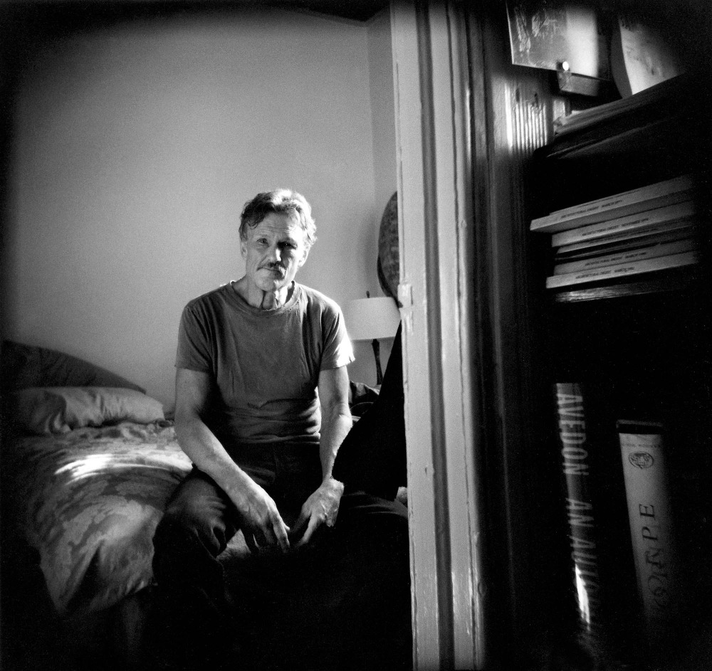 Black and white portrait of a man sitting on a bed in a room with a bookcase, appearing pensive in a medium shot.