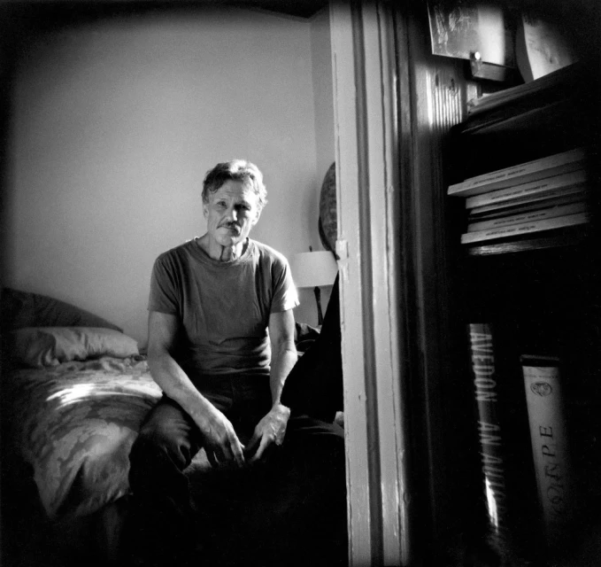 Black and white portrait of a man sitting on a bed in a room with a bookcase, appearing pensive in a medium shot.