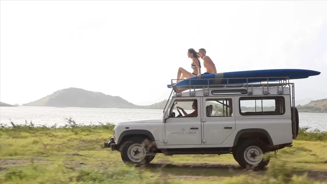 White SUV drives on grassy field by the ocean with a couple sitting on the roof rack with a blue surfboard. 