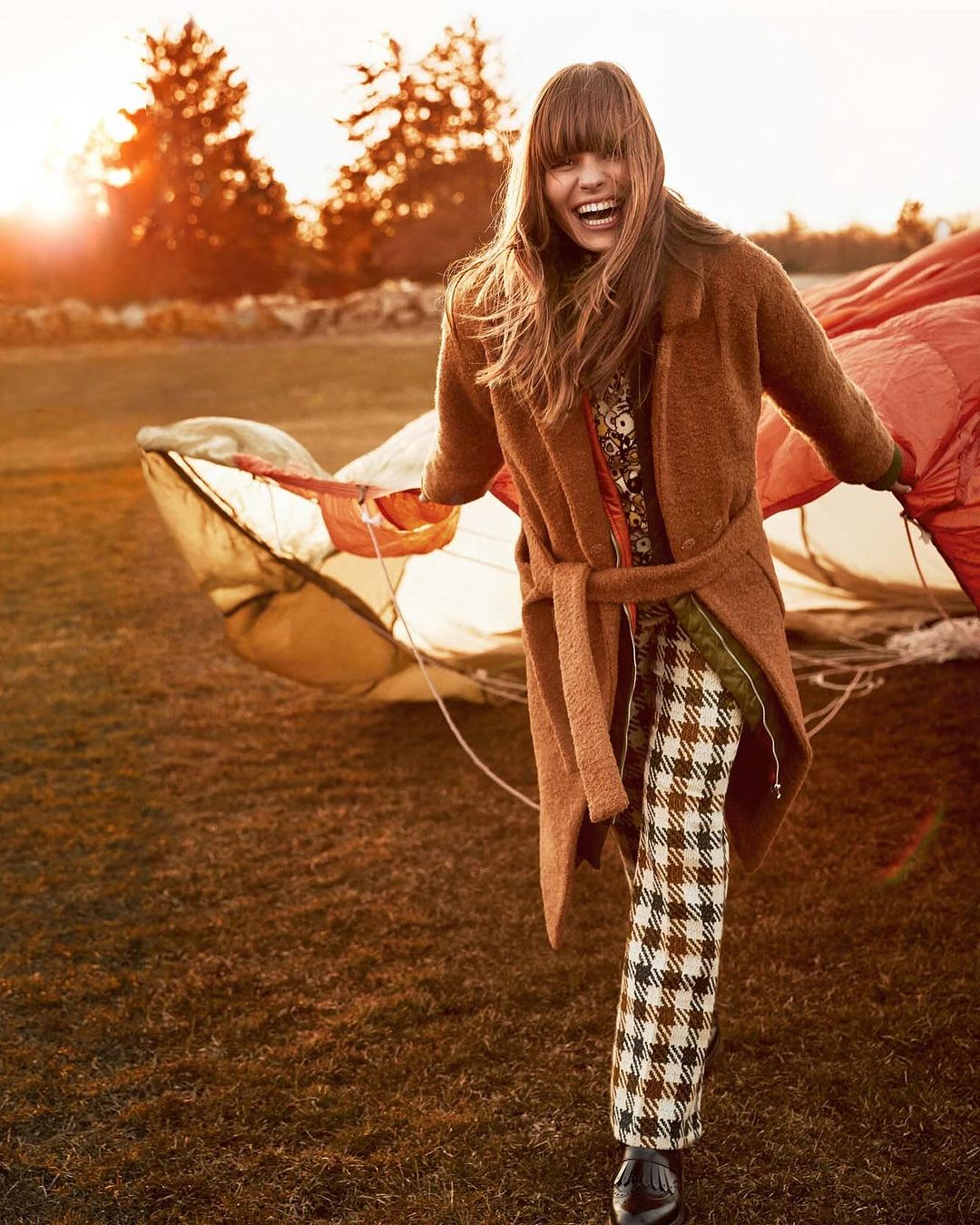 Smiling woman in brown coat and patterned pants stands near partial parachute on grassy field at sunset, fashion editorial.