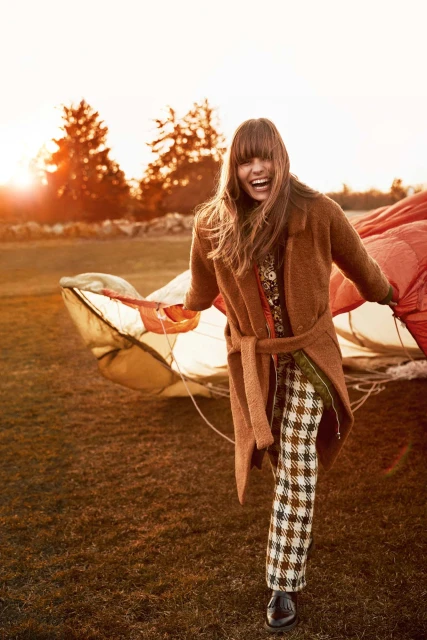 A woman in a brown coat and patterned pants smiles and stands outdoors near a parachute in a field at golden hour.