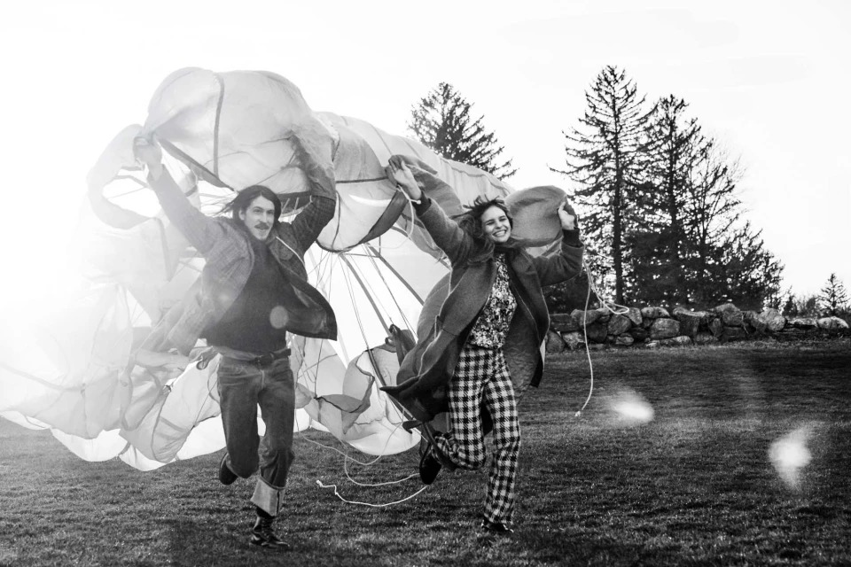 A happy couple in vintage attire run across a grass field, joyfully holding a parachute aloft against a backdrop of trees and sunlight.