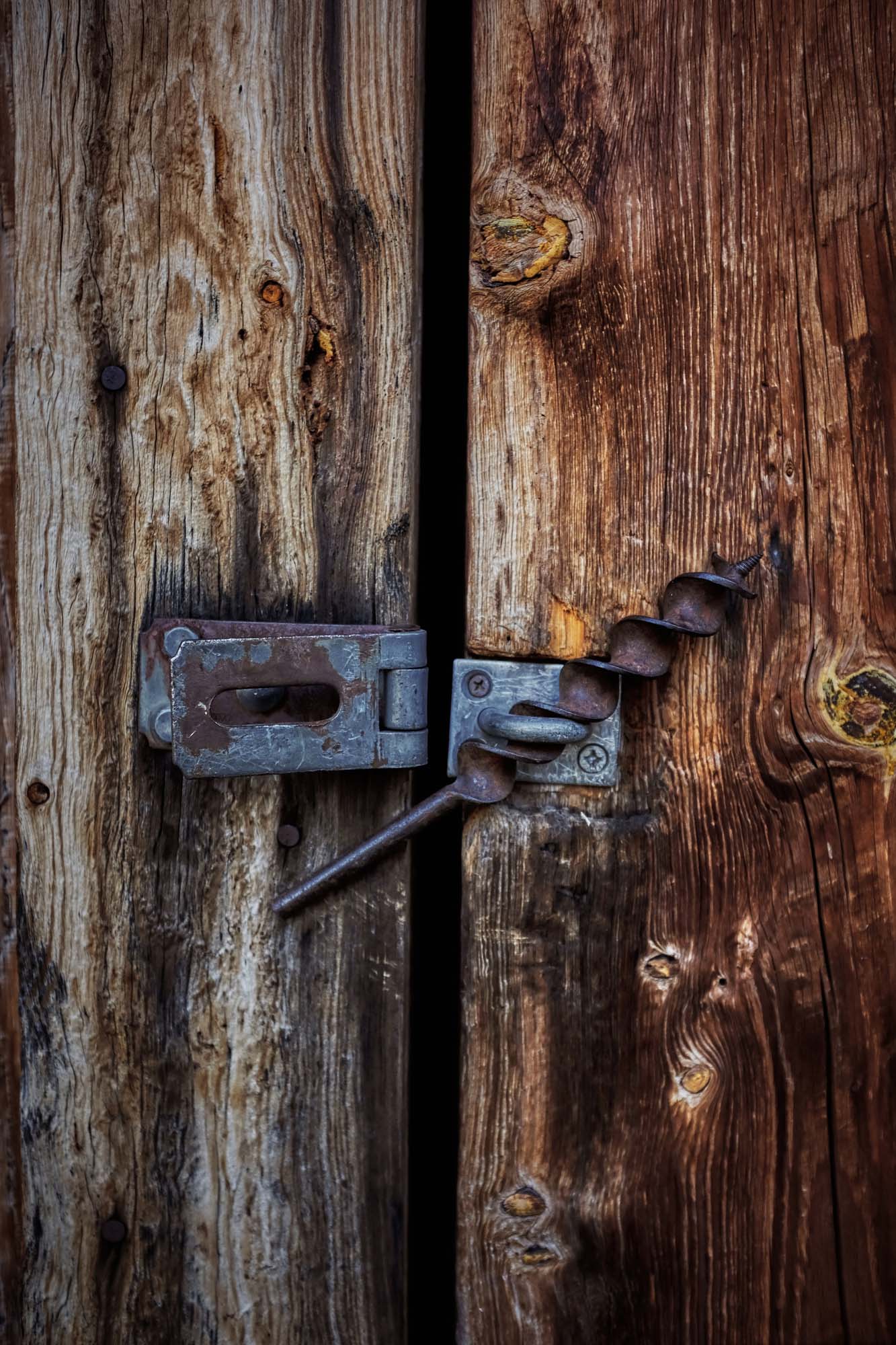 Close-up of a weathered wooden door secured with a rusty metal latch and a vintage-style drill bit serving as a makeshift lock.