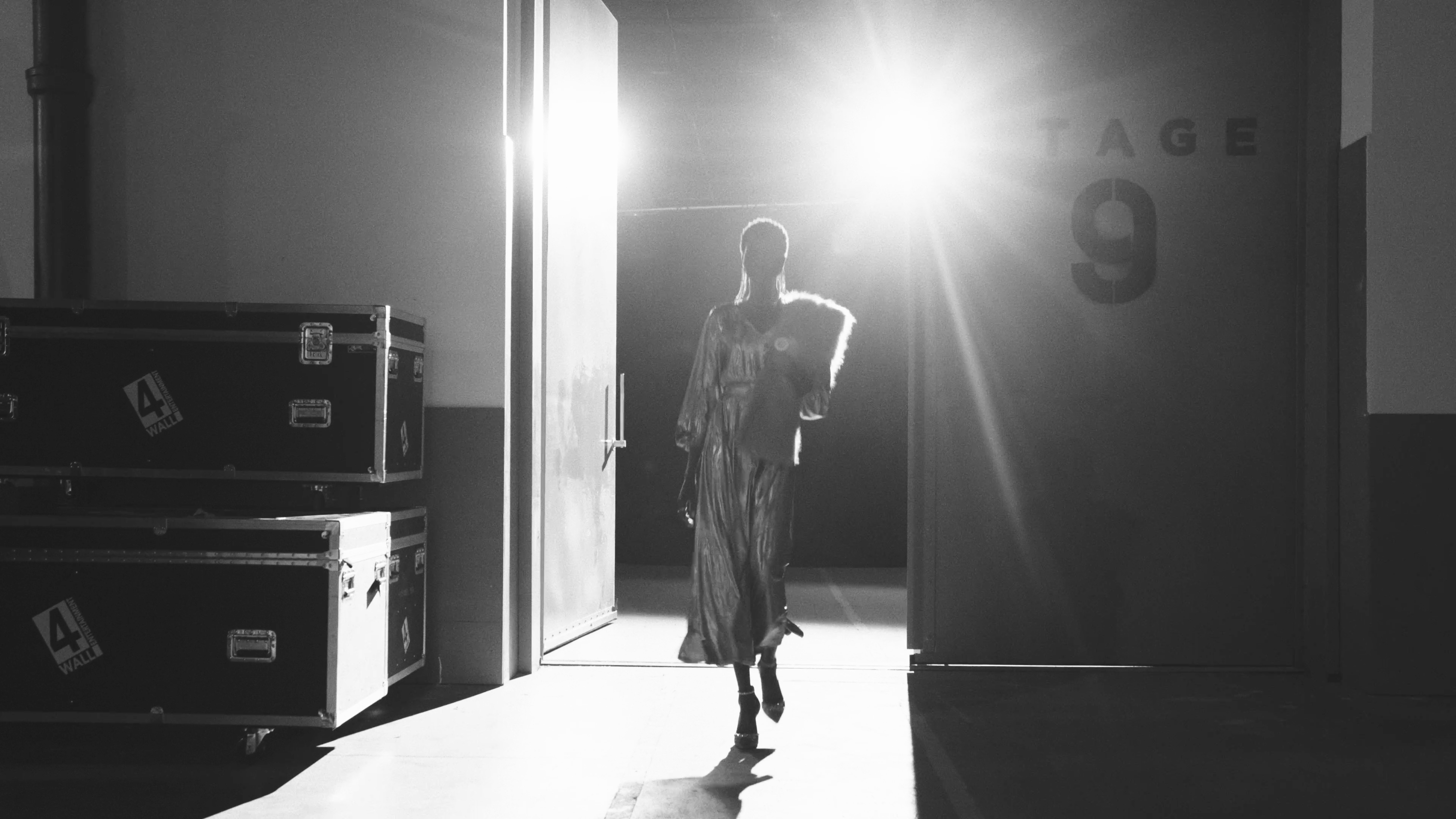 Woman in a shimmering dress walks towards the camera through a backstage doorway with bright backlight, creating a dramatic black and white fashion image.