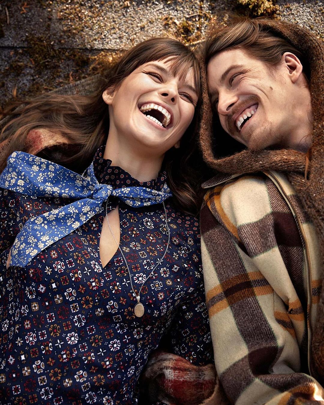 Smiling couple lying on ground with patterned clothing, scarf, and necklace in outdoor setting with neutral background and lighting.