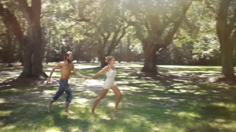 A barefoot man and woman in light dress run hand-in-hand across a green lawn bordered by large trees and bathed in bright sunlight.