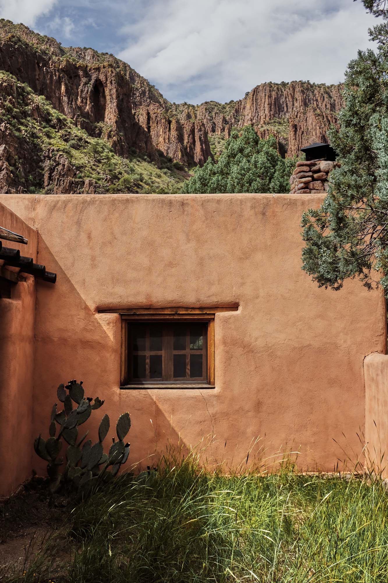 Adobe building with a window sits in front of a mountain range with green plants and shrubs in the foreground under a bright sky.