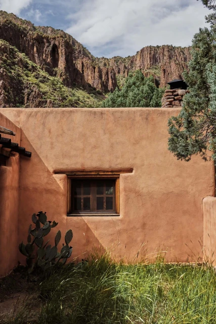 Adobe building with a window sits in front of a mountain range with green plants and shrubs in the foreground under a bright sky.